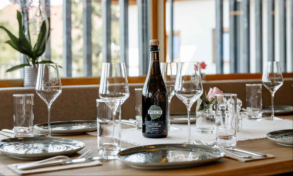 Stilvoll gedeckter Tisch im Buschenschank des Weinguts Pilch mit Weinflasche, Weingläsern und regionalem Ambiente – Genuss mit Blick auf die Südsteiermark.