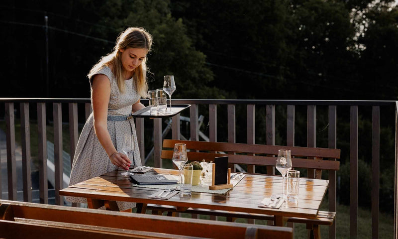 Chefin deckt einen Tisch auf der Panoramaterrasse des Weinguts Pilch für Gäste ein – gemütliches Ambiente mit regionaler Gastfreundschaft und Ausblick über die Südsteiermark.