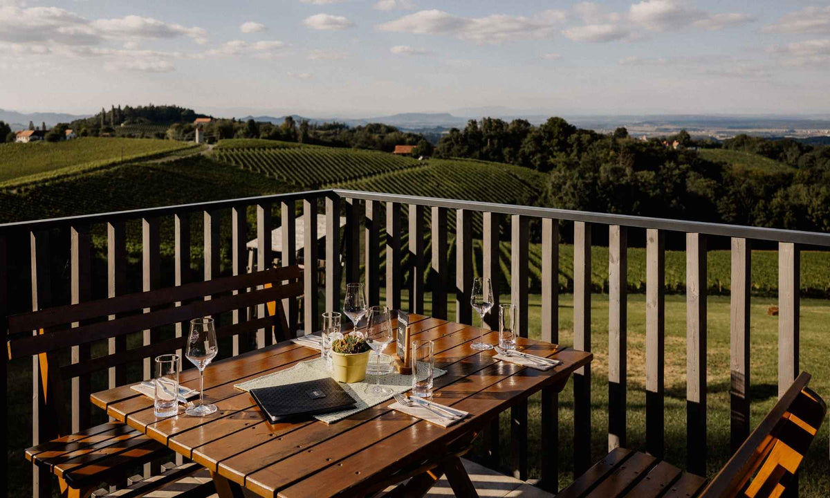 Panoramaterrasse des Weinguts Pilch mit gedecktem Holztisch, Weingläsern und traumhaftem Blick über die Weinberge der Südsteiermark – der perfekte Ort zum Genießen regionaler Spezialitäten und Weine.