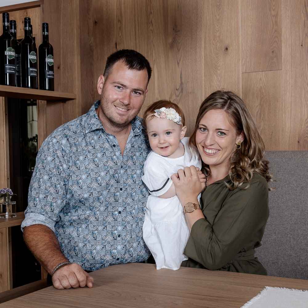 Winzerfamilie Pilch mit ihrem Kind im Verkostungsraum des Weinguts Pilch in der Südsteiermark. Authentisches Familienfoto vor einer warmen Holzkulisse mit Weinflaschen aus eigener Produktion.